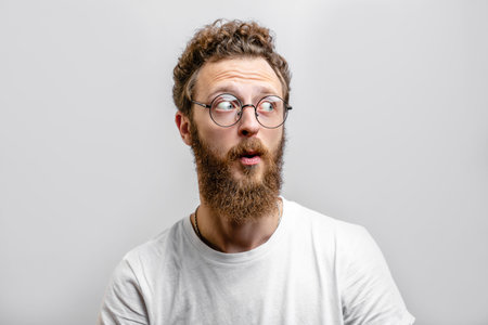 Surprised caucasian man with curly hair and beard stares in amazement aside, dressed in casual white...