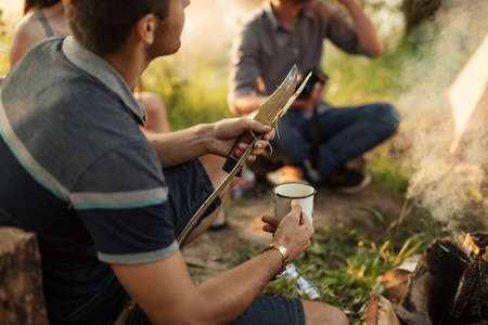 young explore is holding knife and stick and drinking coffee while sitting near the campsite. close up cropped photoの写真素材