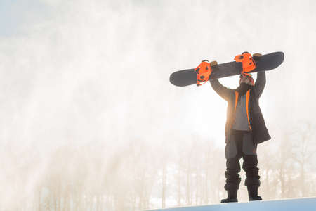 serious snowboarder checking his snowboard before descending. full length photo. copy space. sporty fit man is holding a snowboard over the head and looking at itの写真素材