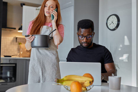 confident african man work on laptop at home while wife is busy in the kitchen, young guy sit at...