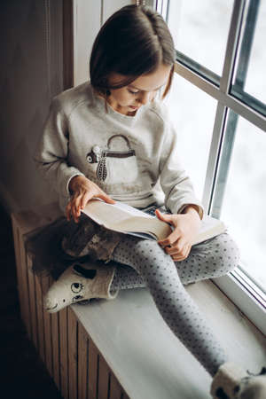 little pretty serious girl reading an interesting book while sitting on windowsill. leisure time. interest, lifestyle. close up shotの写真素材