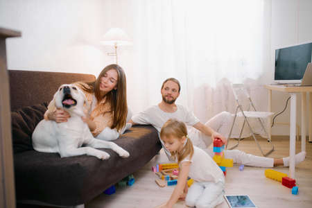 happy caucasian family spending time together at home with lovely friendly pet, domestic animal, white beautiful dog. Young family with children and domestic animal, member of their familyの写真素材