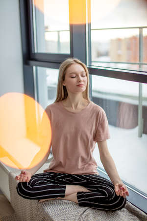 Beautiful daydreamer. Caucasian young woman meditating in lotus pose, keeping eyes closed with smile while sitting at window sill at homeの写真素材