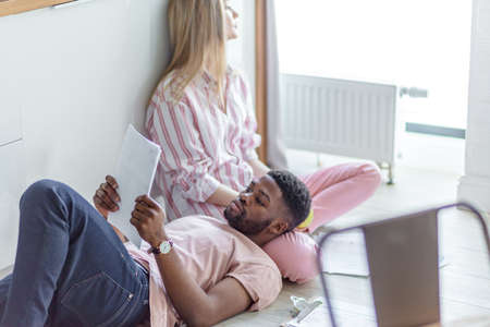 African man reading papers while resting on knees of his blonde long-haired caucasian girlfriend, sitting in sleepwear on the floor at homeの写真素材