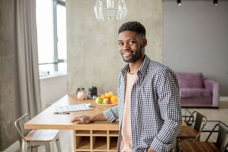 Happy young African bearded man in casual clothes looking at camera, posing over modern home interior.の写真素材