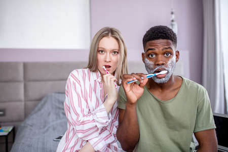 Mixed race couple brushing teeth in morning, looking at camera with amazement.の写真素材