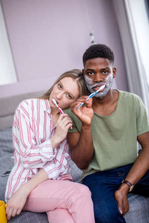 Attractive multiethnic couple brushing their teeth in the morning at home, african man having shaving cream on his cheeksの写真素材