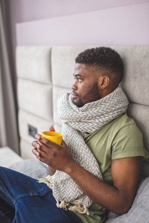 Young African businessman being upset and distempered with his illness, wrapped in knitted thick...