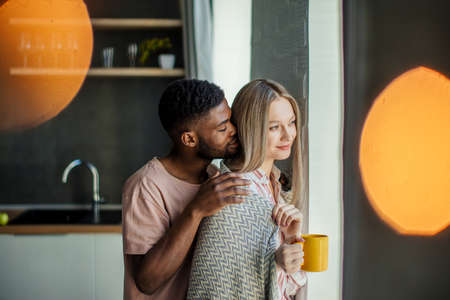Handsome african guy kissing his caucasian girlfriend on the neck from backside, standing at home in the kitchen room with modern interiorの写真素材