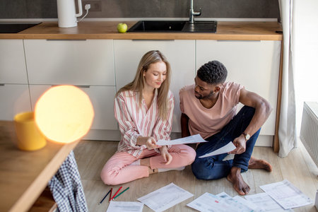 Young mixed-race couple of diverse students in casual wear, sitting together on kitchen floor at...
