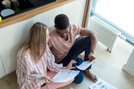 Young multi-ethnic couple sit close at the kitchen floor, planning their joint summer vacations,...