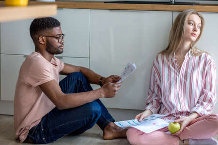 Serious african man comforts his depressed caucasian girlfriend, being upset with bad news from letter she recently had got. Couple sitting on floor, sorrowful girl in casual wear looks asideの写真素材