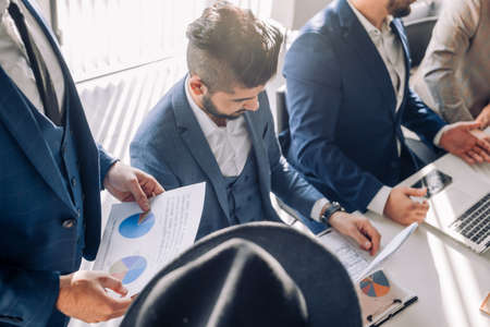 Respectable Indian employee dressed in formal suit and hat planning company costs, discussing financial strategy or marketing research during meeting at desk in office.の写真素材