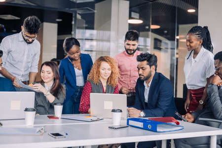 Creative multiracial business team discussing project, standing around workplace, while two co-workers with portable computers looking for needed information, sharing results to the workersの写真素材