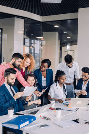 Searching of new insights and brilliant ideas. Mixed-race group of nine diverse people browsing net and looking through documents while sitting at the table in officeの写真素材