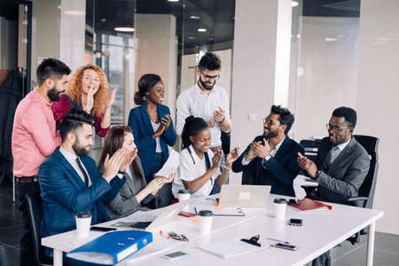 Mixed-race group of nine diverse happy people standing arround the table with colleagues seated , applauding team achievementsの写真素材