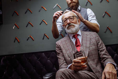 Barber making male haircut. Hairdresser cutting hair of his rich client, dressed in expensive stylish man s suit of clothes and holding a glass of alcoholic beverageの写真素材