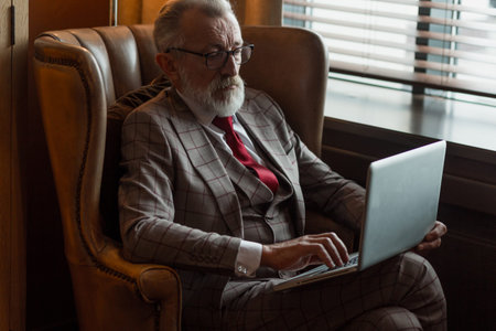Fashionable focused elderly male writer wearing glasses and elegant clothes working on laptop, close...