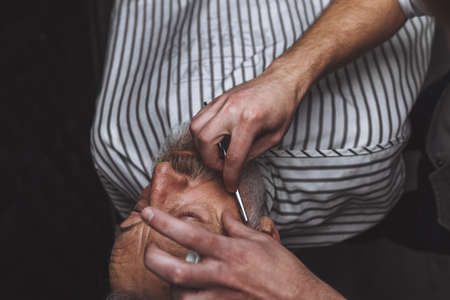 Respectable senior caucasian man visiting hairstylist in barber shop for shaving and making new design for his grey-haired beardの写真素材
