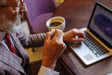 High angle view of a confident old European businessman wearing spectacles, looking at newspaper laptop holding a cup of coffee while sitting at a table with laptop in a cafeの写真素材