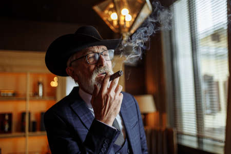 Stylish old-aged dandy in wide brimmed hat and rich dark blue mens suit smoking cigar indoor, standing near the bar counter with alcohol drinks.の写真素材