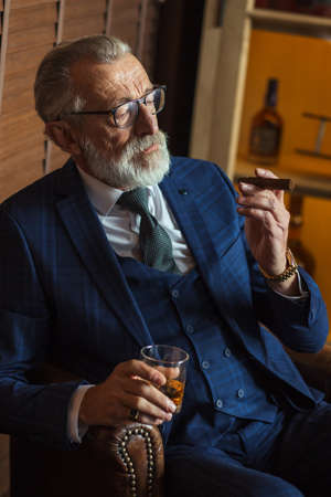Senior cute business man with cigar and whisky. Gray hair and beard wearing blue tailored suit and tie, relaxing at fashionable restaurant loungeの写真素材