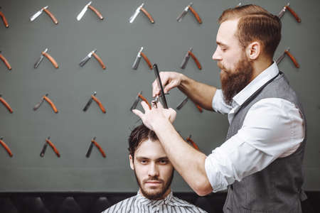 Working process in modern barbershop. Male hairdresser serving client, making haircut using metal scissors and comb, side view.の写真素材