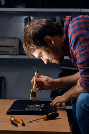 handsome man using brush to clean the laptop from the dust, close up side view photo. profession,...