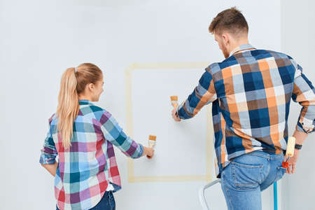 Caucasian young couple painting walls in new apartment with their own hands using paint rollers and paintbrush on wall.の写真素材