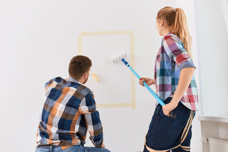Happy smiling young couple painting interior white walls renovating their new home together using rollers on long handleの写真素材