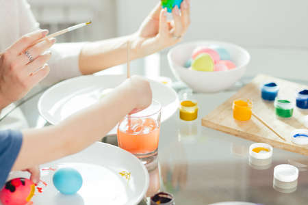 Home made Easter decorations and little helper. Selective focus of small hands of a boy covering boiled eggs with colourful paints on white plateの写真素材