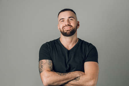Young strong happy bearded man with arms crossed isolated on gray background. close up shot. sport, fitness, health, strength, wellnessの写真素材