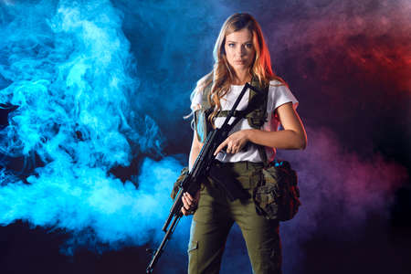Young blonde female snipper in military outfit with assault rifle in studio on smoky dark background. Women in military service concept.の写真素材