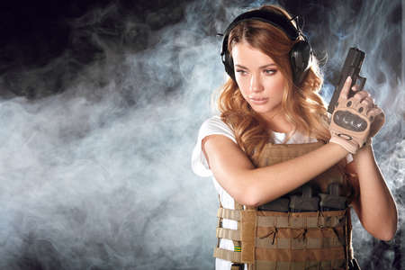 Armed beautiful blonde woman wearing protective headphones and plate carrier, shoots with gun at a target in the darkness with smoke cloudsの写真素材