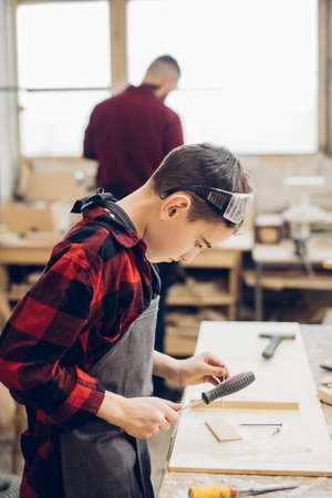 Little male helper working with his male teacher building wooden kit during woodworking classes. 9 year old boy in working apron and protective glasses using screwdriver.の写真素材