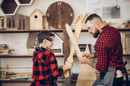 Family relations, fatherhood, hobby, carpentry, woodwork concept - Father and son playing knights with wooden DIY swords at carpenter workshopの写真素材