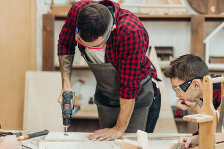 Little teen boy helps his father, working as woodworker, to drill holes in a wooden plank. Close-up of young male carpenter in protective eyewear teaching his son to work with wood in his workshop.の写真素材