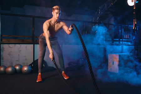 Handsome muscular champion man is doing battle rope exercise while working out in dark gym,...