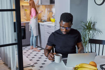 confident african man work on laptop at home while wife is busy in the kitchen, young guy sit at table concentrated on freelance work, redhead lady in apron does household choresの写真素材