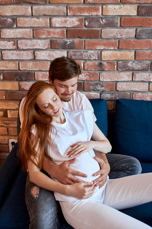 careful responsive man stroke pregnant belly of wife, lovely couple have rest on sofa in living room...