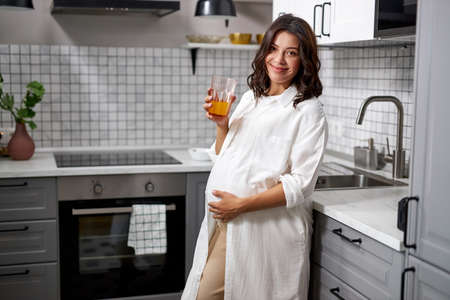 portrait of smiling pregnant woman with cup of juice in hands, adorable lady look at camera, hold belly. in kitchenの写真素材