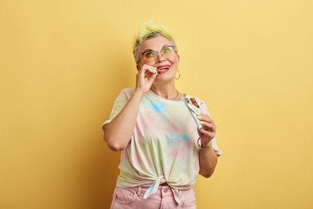 young glamour old woman enjoying eating chocolate isolated over yellow wall background, old woman...