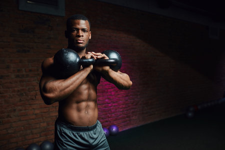 Young ripped African male athlete with perfect well-shaped body posing with dumbbells on brick wall...