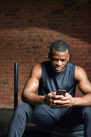 Portrait of african athlete man checking his smart phone while resting after training in gym.