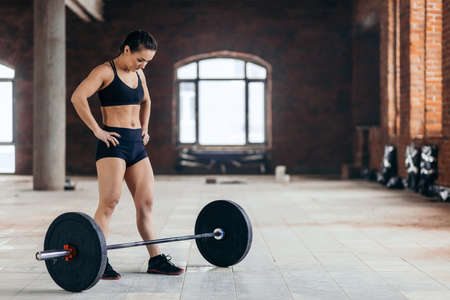 tired beautiful young woman with hands on the hips resting after workout with barbell. Fitness, sports, cross fit. copy space. full length photo.の写真素材