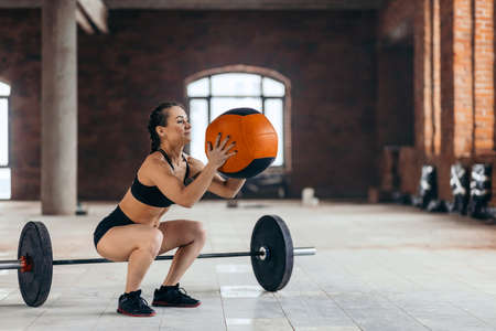 well-built motivated girl doing squat exercises with fitness ball. barbell on the floor on the background of the photo, combination of croos fit exercisesの写真素材
