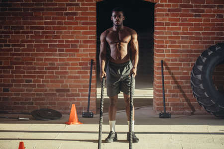 African muscular shirtless man is concentrated on the exercise with battle rope requiring increased effort and strength, standing at half squatting over brick backgroundの写真素材