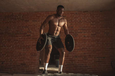 African american crossfit sportsmen holding barbell plates as weighting in both hands making jumps on big rubber wheel, lying on the floor of street local gym.の写真素材
