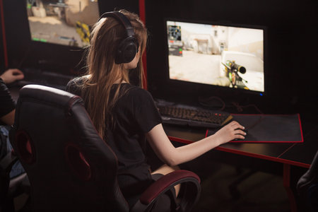 Rear view of female gamer girl with headset sits in a dark room behind a computer with a raw of...