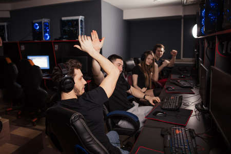 Enthusiastic e-sport gamers. Joyful young people sitting at powerful computers in gaming centre and raising hands triumphantly, celebrating victory in game.の写真素材